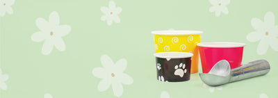 Assorted colorful ice cream cups, including yellow with white spirals, black with white paw prints, and a plain red cup, arranged next to a metallic ice cream scoop on a light green background with white flower patterns